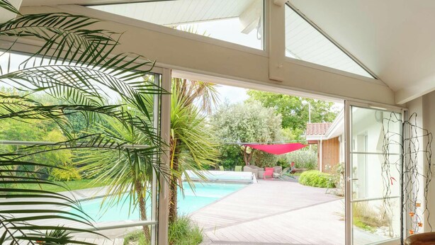 Ein modernes Haus mit Glastür, die zu einem Poolbereich führt, umgeben von viel Grün.