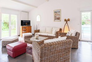 A living room with wicker furniture, a red ottoman, a television, and a painting on the wall. - One-story villa with pool - 8 guests (Biscarrosse)