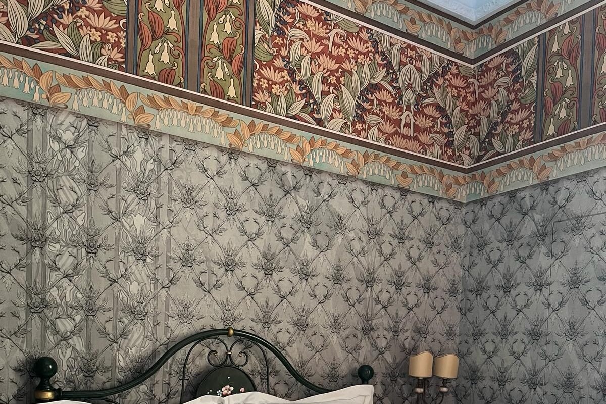 Un letto ben rifatto con lenzuola bianche e verdi, una testiera in metallo scuro e carta da parati con motivi floreali.