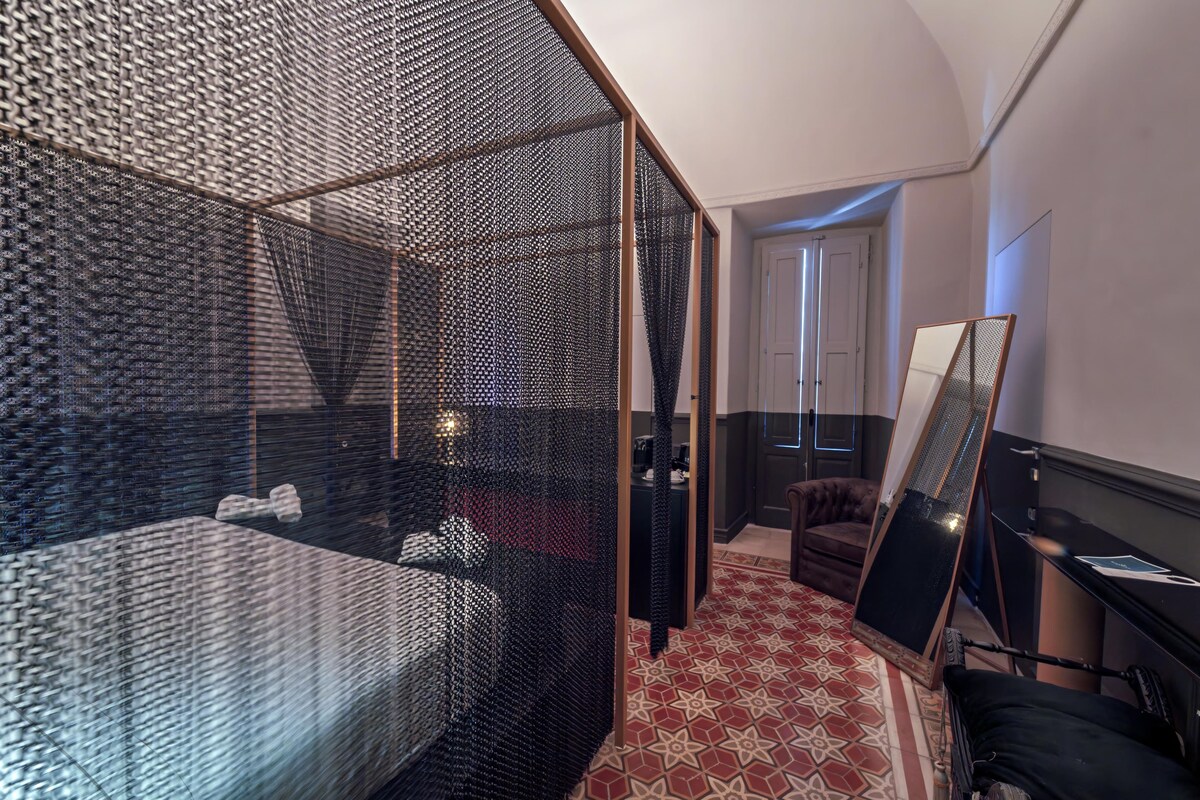 Una camera d'albergo moderna con un letto grande, un'area salotto con una poltrona, una scrivania e un ampio specchio.