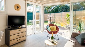 A modern living room with a TV, a sofa, a dining area, and a view of the backyard. - Charming house in Navarrosse - 6 guests (Biscarrosse)