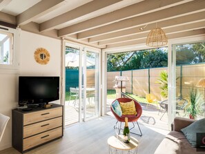 A modern living room with a TV, a sofa, a dining area, and a view of the backyard. - Charming house in Navarrosse - 6 guests (Biscarrosse)