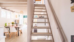A modern interior with a wooden staircase, a dining area with white chairs, and a kitchen visible through an open door. - Charming house in Navarrosse - 6 guests (Biscarrosse)