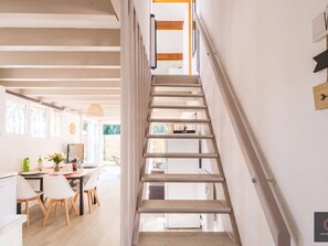 A modern interior with a wooden staircase, a dining area with white chairs, and a kitchen visible through an open door. - Charming house in Navarrosse - 6 guests (Biscarrosse)
