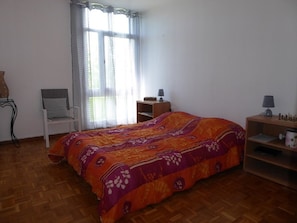 A bedroom with a bed, a chair, a nightstand, and a window with curtains. - Charming apartment in Biscarrosse city center (Biscarrosse)