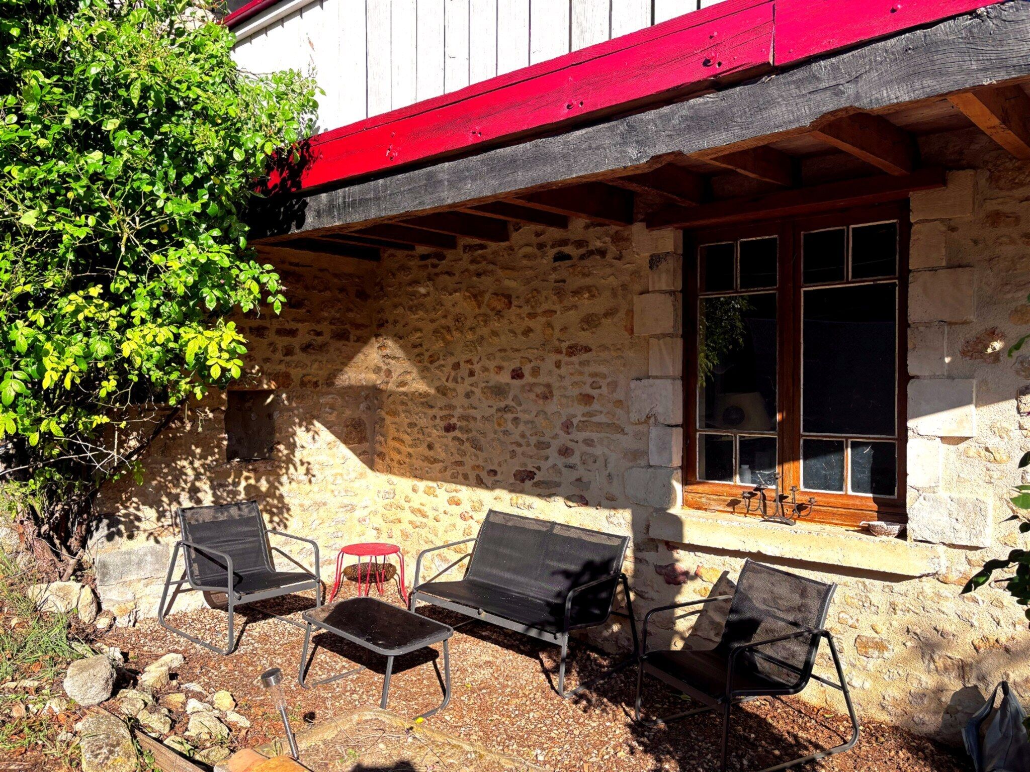 A stone building with a red roof, a window with a view, and three black chairs outside.