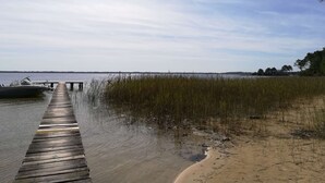 A wooden pier extending into calm water with a sandy beach and reeds on the right.