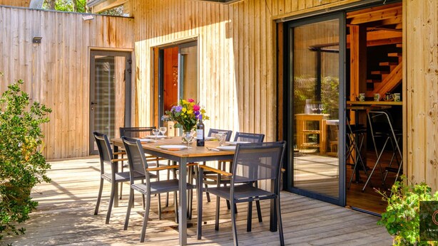 A wooden deck with a table and chairs, a glass door leading to an interior space, and a small garden area.
