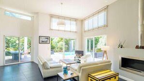 A spacious living area with a sofa, coffee table, and a fireplace. There are large windows and a view of the outdoors.