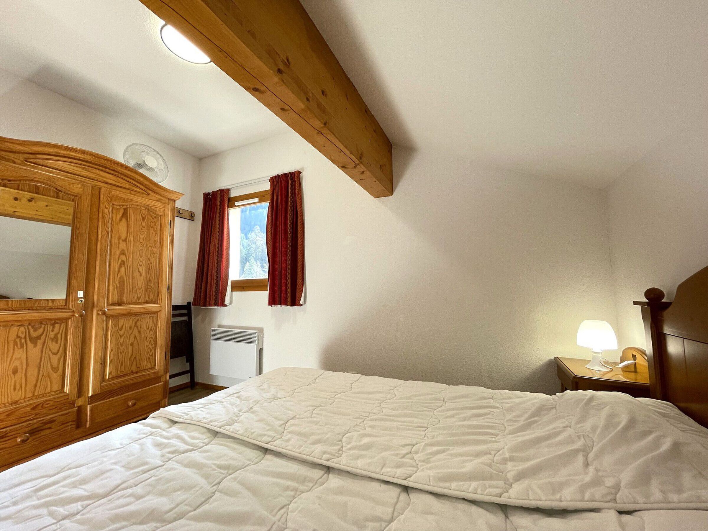 A bedroom with a wooden wardrobe, a bed with a white quilt, a window with red curtains, a bedside lamp, and a radiator.