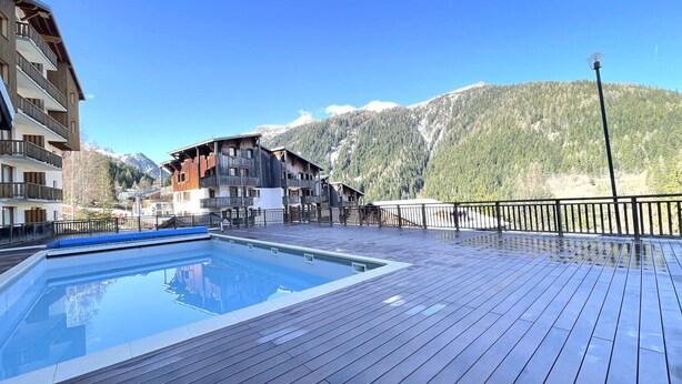 A wooden deck with a pool, a mountain backdrop, and a building with balconies. - Comfortable duplex, slope view - 6 guests (Valfréjus)