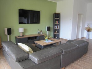 A living room with a sectional sofa, a coffee table, a TV mounted on the wall, a bookshelf, and a speaker. - After stand UP paddling, barbecue on the terrace and enjoy the sunsets (Klein Köris)