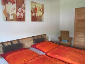 Una cama con ropa de cama roja y naranja, un cabecero de mimbre, una silla y dos cuadros colgados en la pared.
