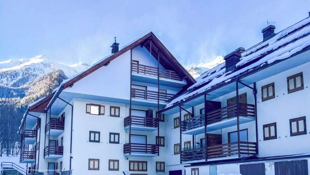 A multi-story building with balconies, a mountain backdrop, and snow-covered roofs. - Casa Castoro - Nel cuore della Val di Peio (Peio Fonti)