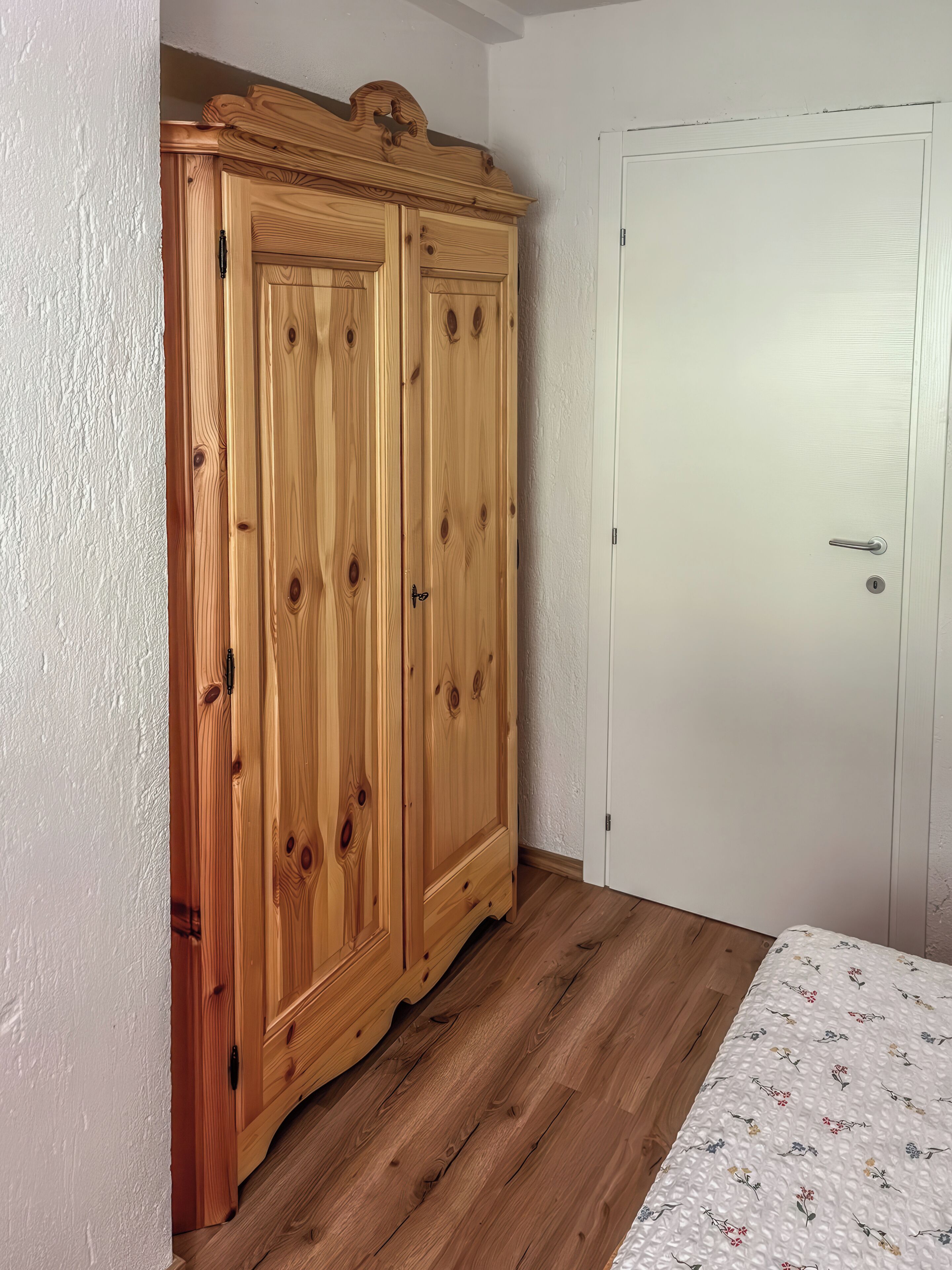 A bedroom with a wooden wardrobe, a white door, and a floral-patterned bedspread.
