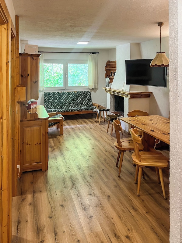 A wooden-floored living area with a sofa, a dining table with chairs, a TV mounted on the wall, and a window with curtains. - Casa Castoro - Nel cuore della Val di Peio (Peio Fonti)