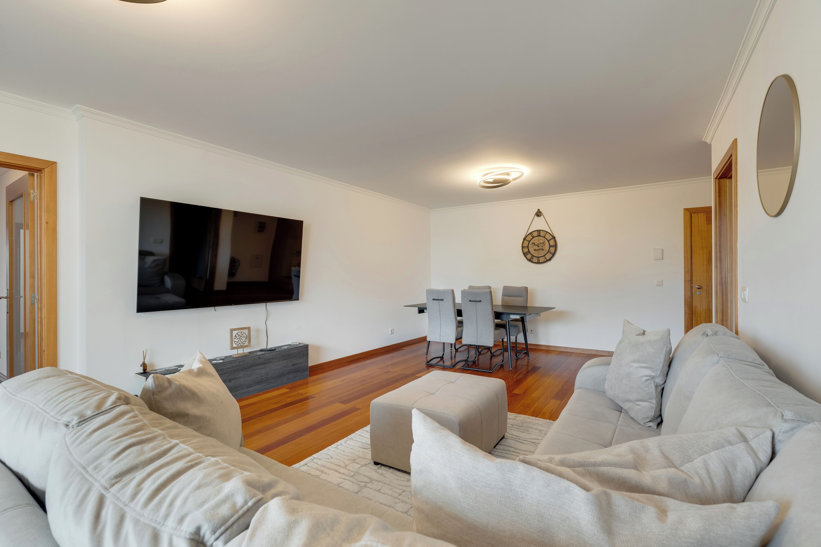 A modern living room with a flat-screen TV, a dining area with a table and chairs, and a large mirror on the wall.