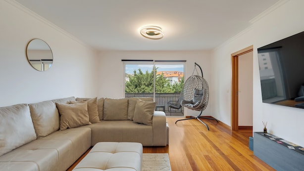 A modern living room with a sectional sofa, a hanging chair, a flat-screen TV, and a large window with a view of buildings and trees. - Sun Flower Apartment (Santa Cruz)