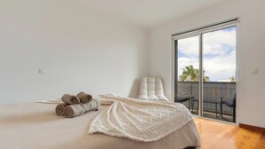 A bedroom with a bed, a white chair, and a balcony with a view. - Sun Flower Apartment (Santa Cruz)