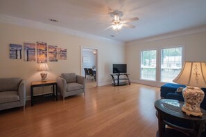 A spacious living room with hardwood flooring, a ceiling fan, a television on a stand, and a dining area visible through an open door.