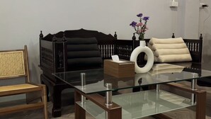 Un salon moderne avec une table basse en verre, un canapé noir, un fauteuil en rotin et un vase rempli de fleurs.