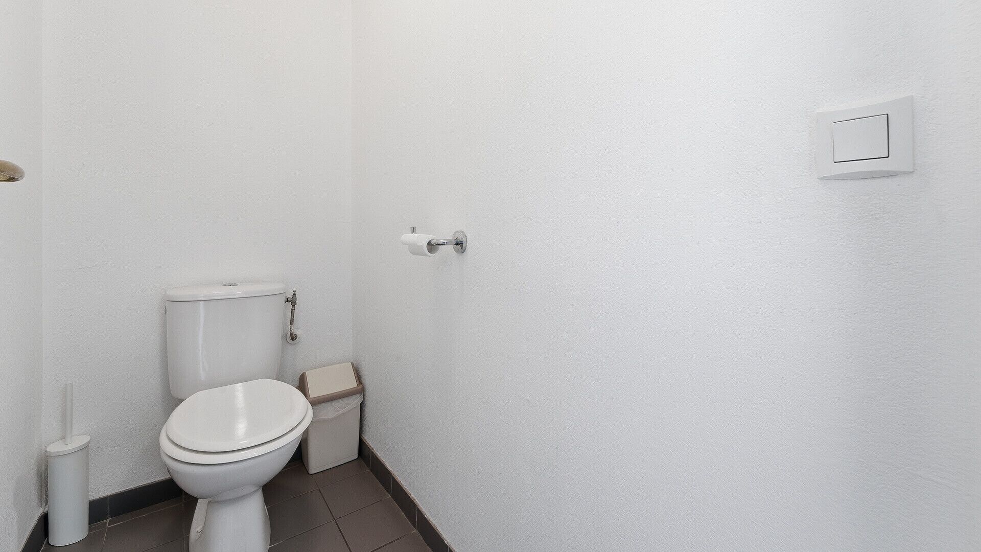Une petite salle de bain aux murs blancs, équipée de toilettes, d’une poubelle et d’une poignée de chasse d’eau fixée au mur.