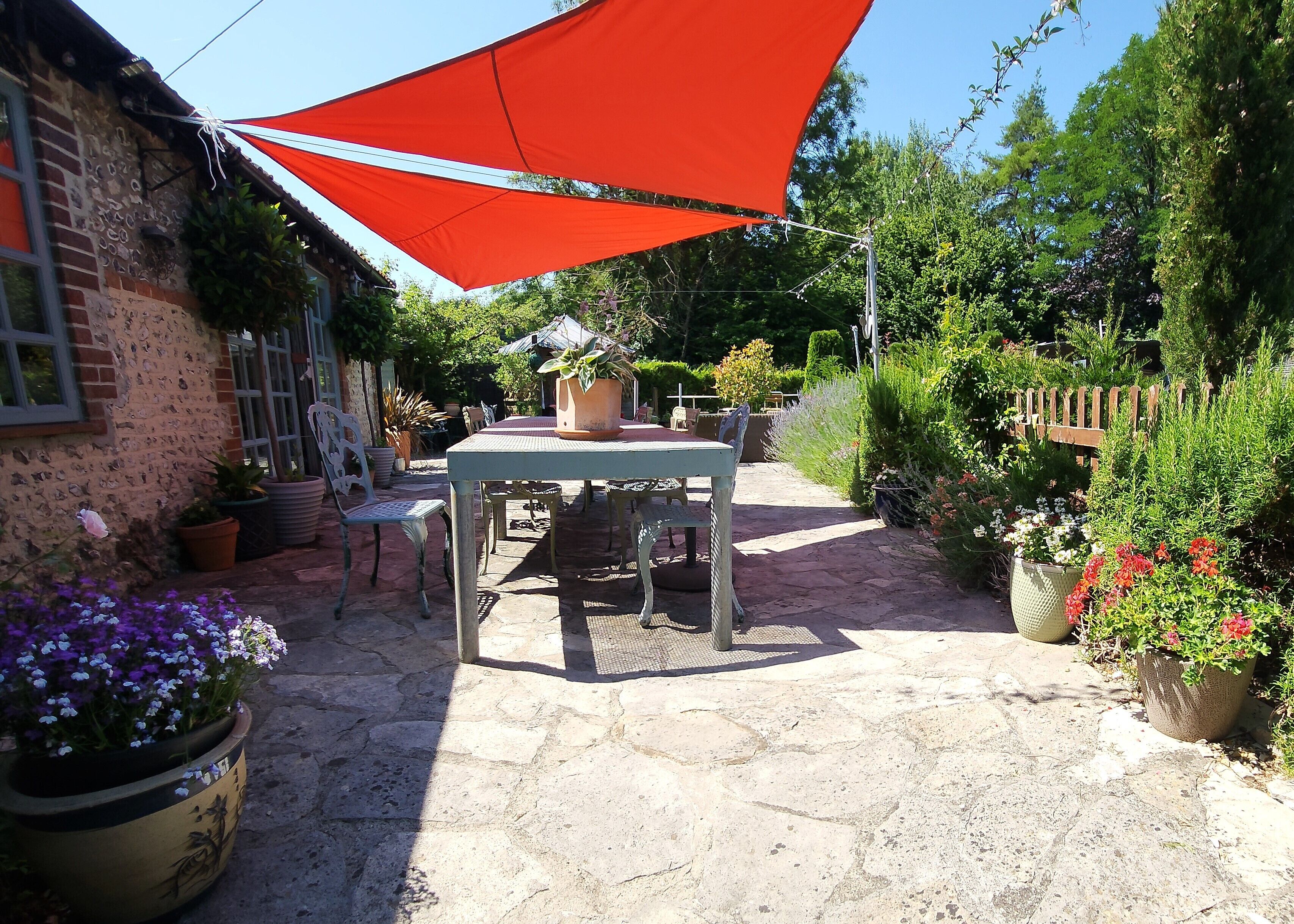 Eine Terrasse mit Tisch und Stühlen, umgeben von Kübelpflanzen und einem roten Sonnensegel.