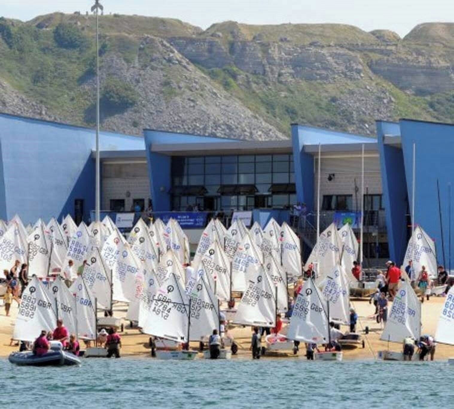 Un puerto deportivo con numerosos veleros amarrados, un edificio con paredes exteriores azules y un telón de fondo montañoso.