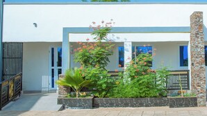 A building with a white exterior, a stone pillar, and a garden with various plants and flowers. - Aureza Paradise Hotel (Tamale)