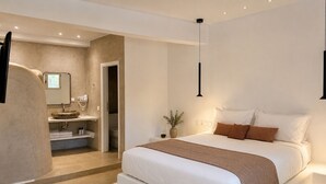A bedroom with a large bed, a bedside table, a nightstand, a mirror, a hanging light, and a bathroom with a sink and toilet.