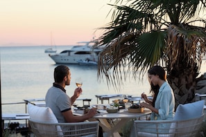 Una mesa de comedor con comida y bebidas, dos personas sentadas, y una vista al mar con yates al fondo.
