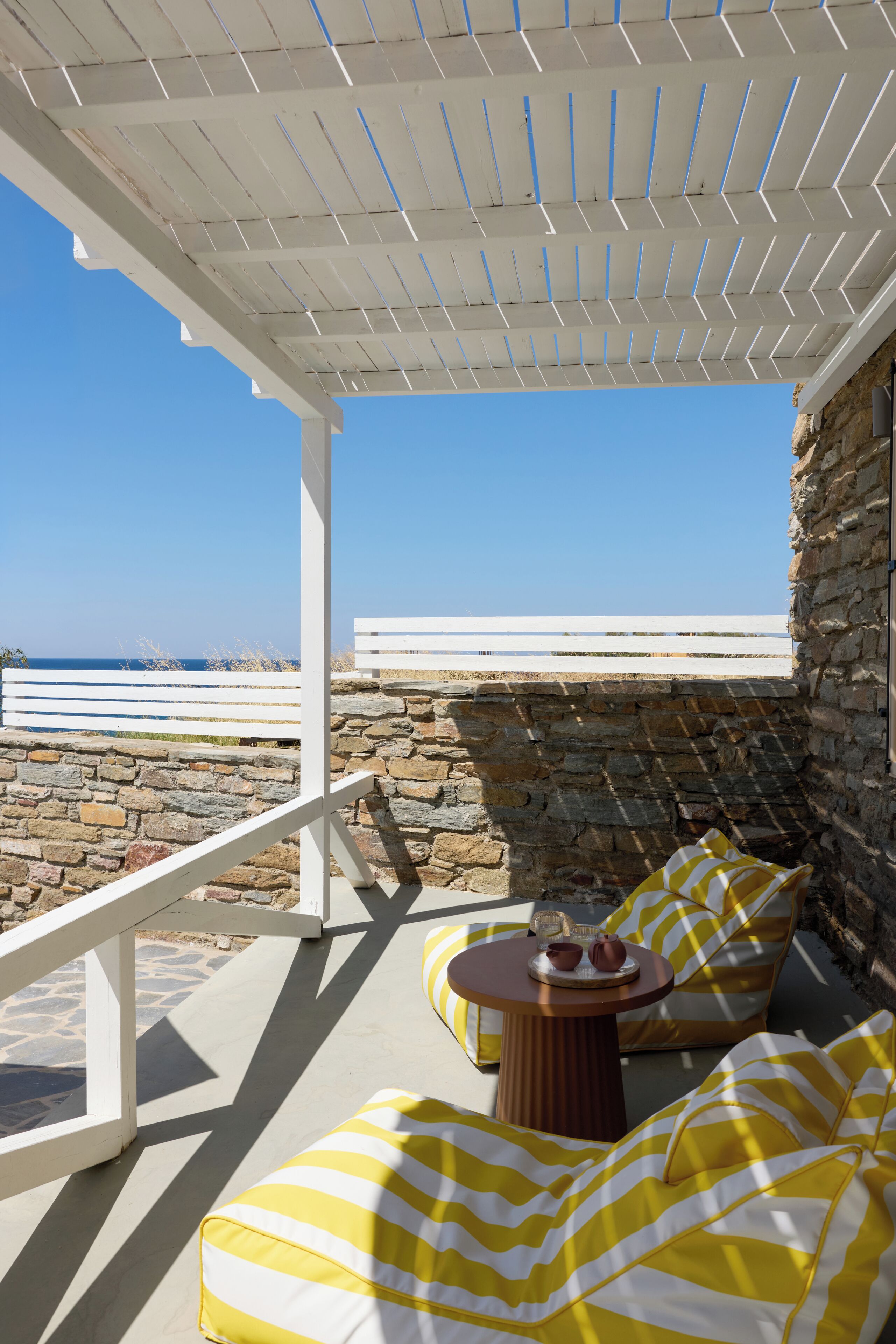 A covered outdoor seating area with stone walls, a view of the sea, and a white railing.