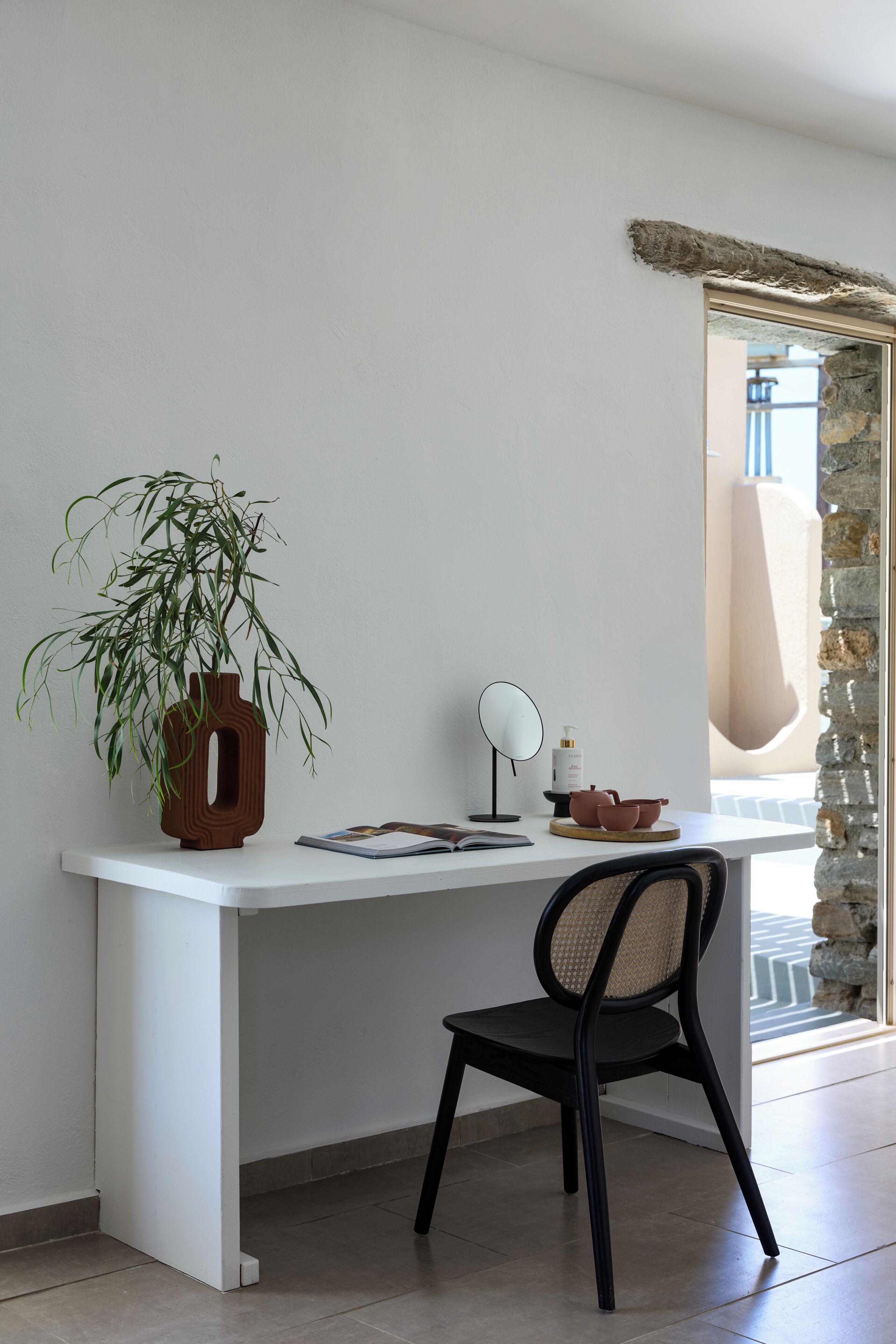 A modern workspace with a white desk, a black chair, a potted plant, a mirror, and a bottle of lotion.