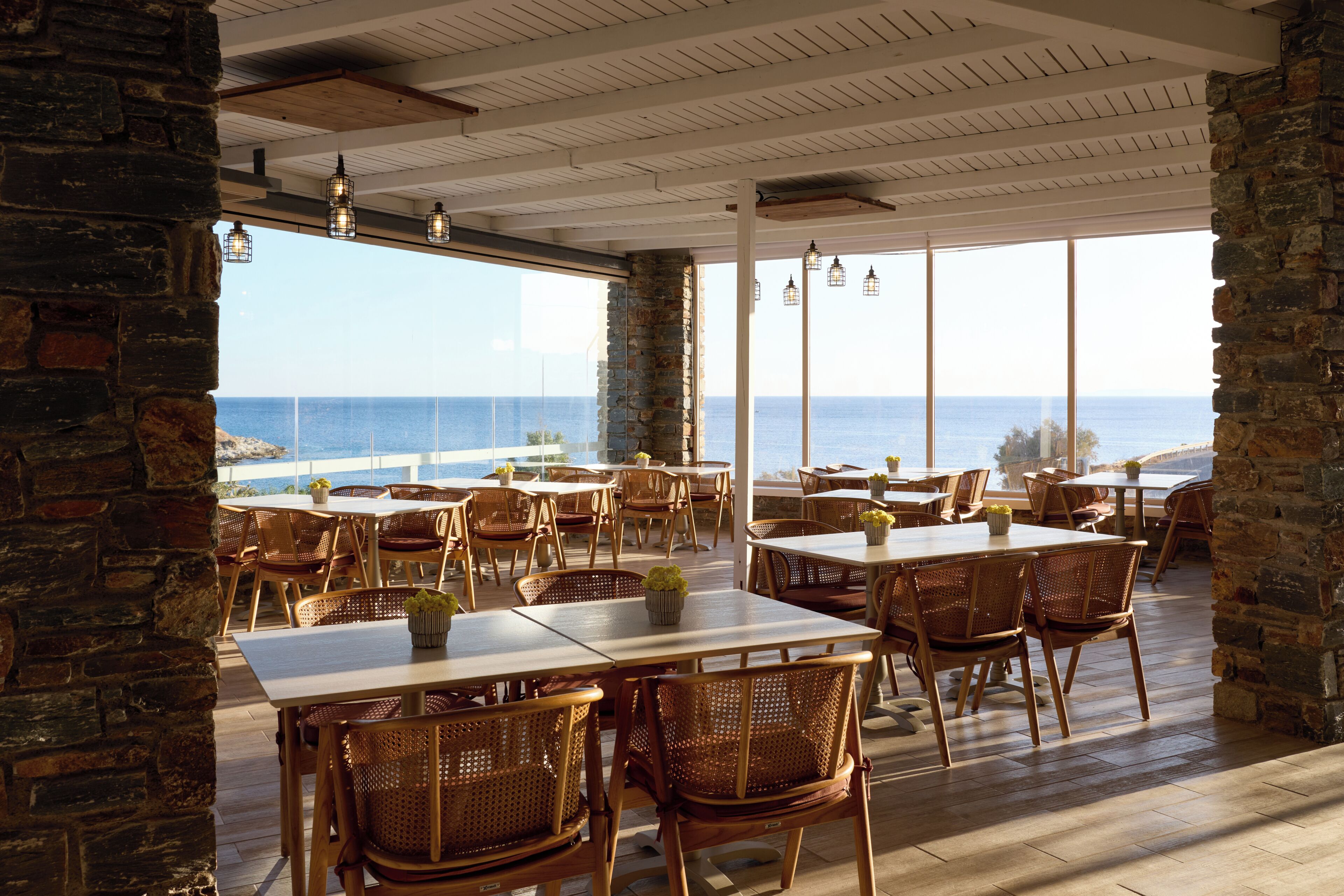 A restaurant with tables and chairs, large windows, and a view of the sea.