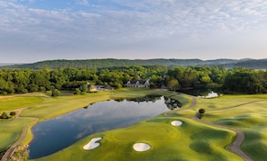 Ein Golfplatz mit Teich, Sandbunkern und einem Clubhaus, umgeben von Bäumen und Hügeln.