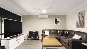 A living room with a flat-screen TV, a brown leather couch, a wooden coffee table, a ceiling fan, and a mounted air conditioner. - Modern 4BR Muswellbrook Stay (Muswellbrook)
