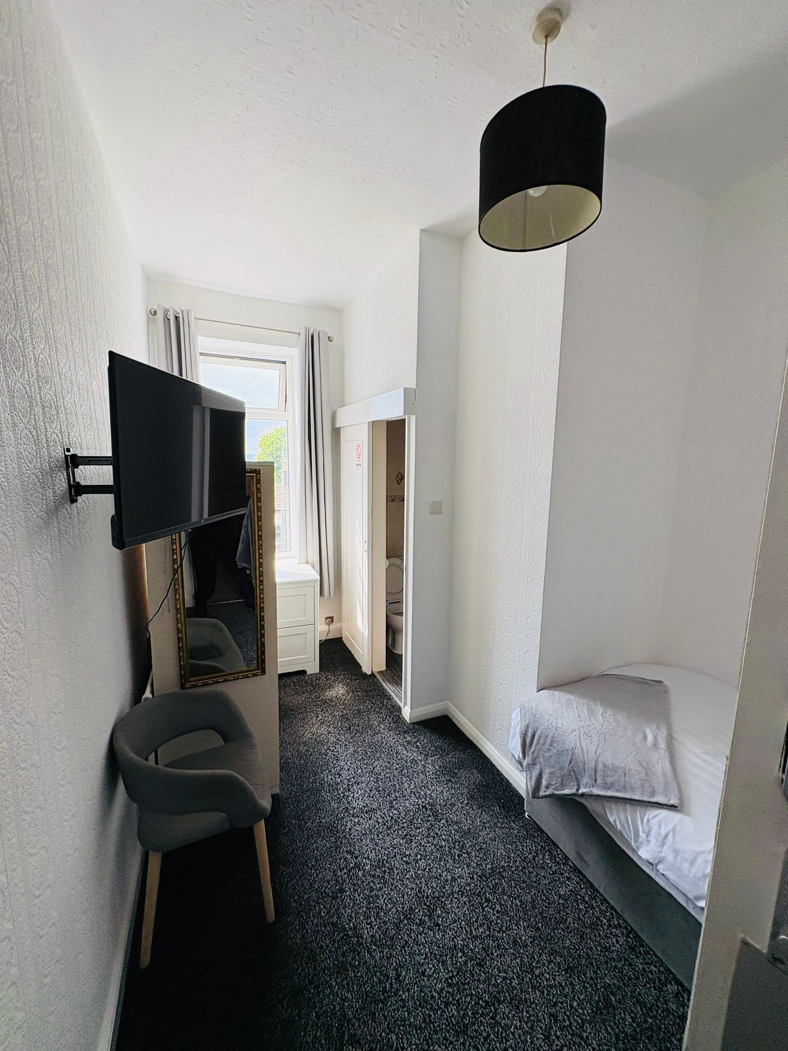A compact hotel room with a bed, a small desk with a chair, a television mounted on the wall, and a ceiling light fixture.