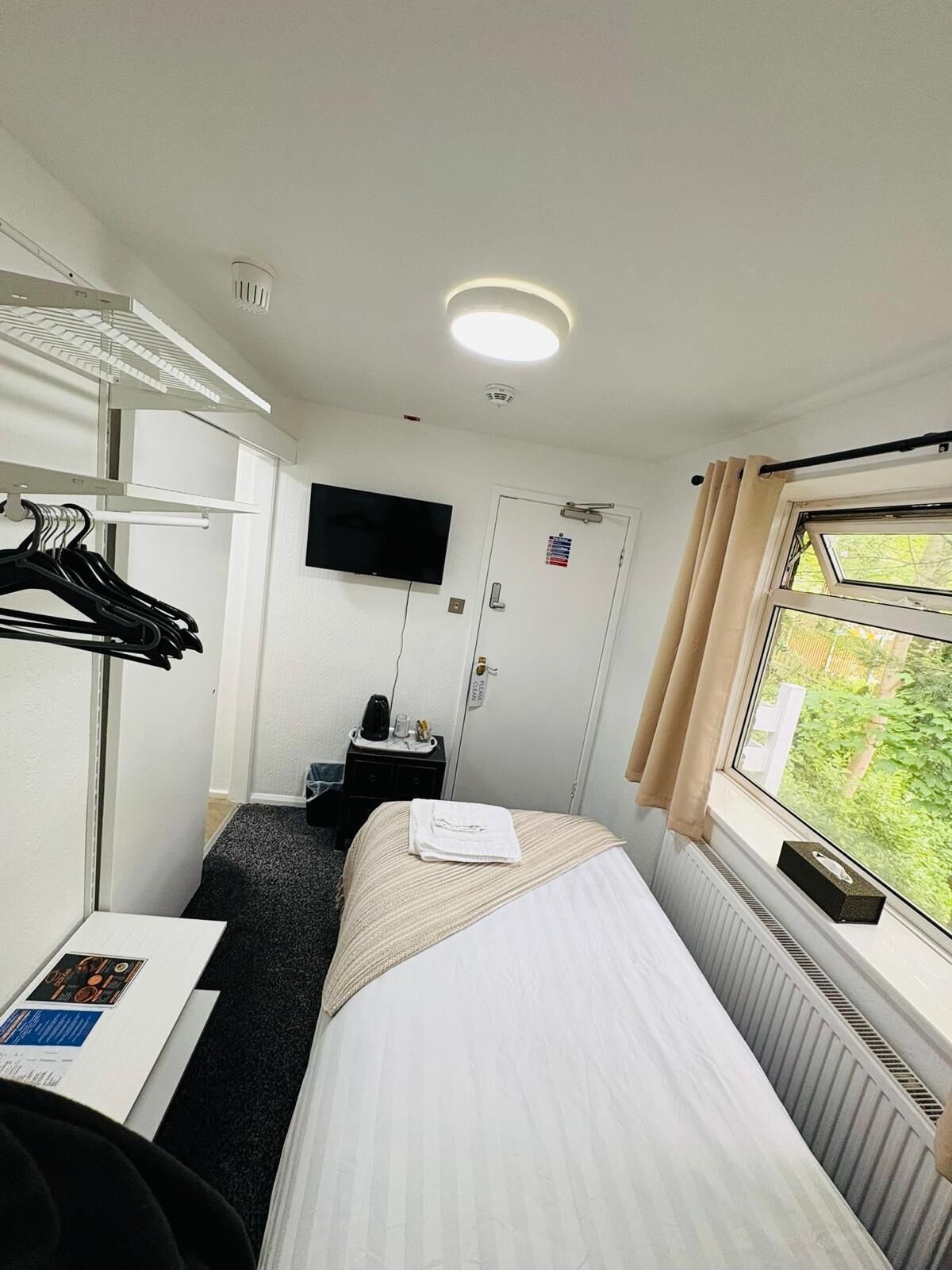 Habitación de hotel con cama, televisión montada en la pared, perchero, escritorio con computadora y ventana con cortinas.