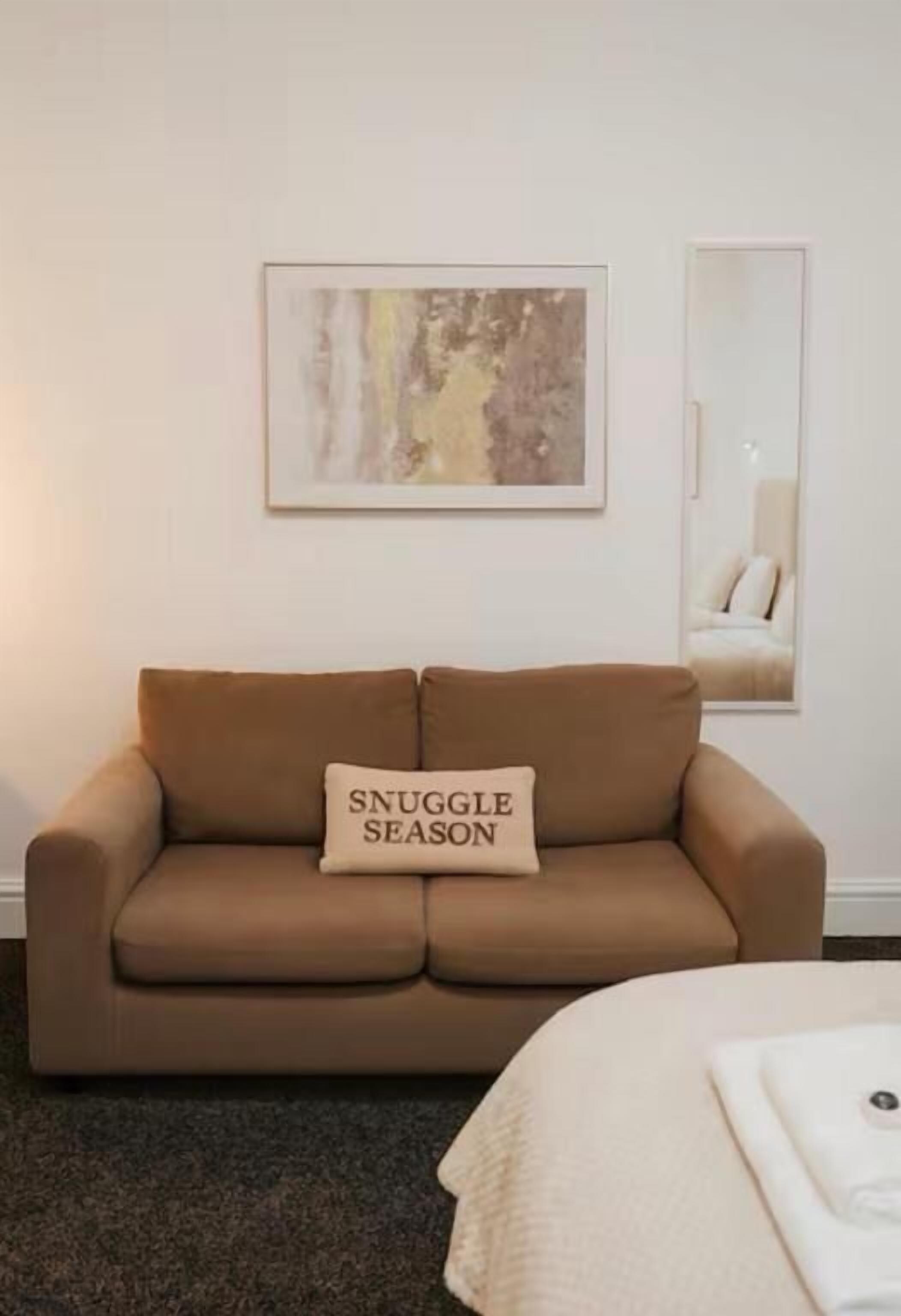 Un sofá marrón con un cojín que dice 'SNUGGLE SEASON' y un cuadro abstracto enmarcado en la pared.