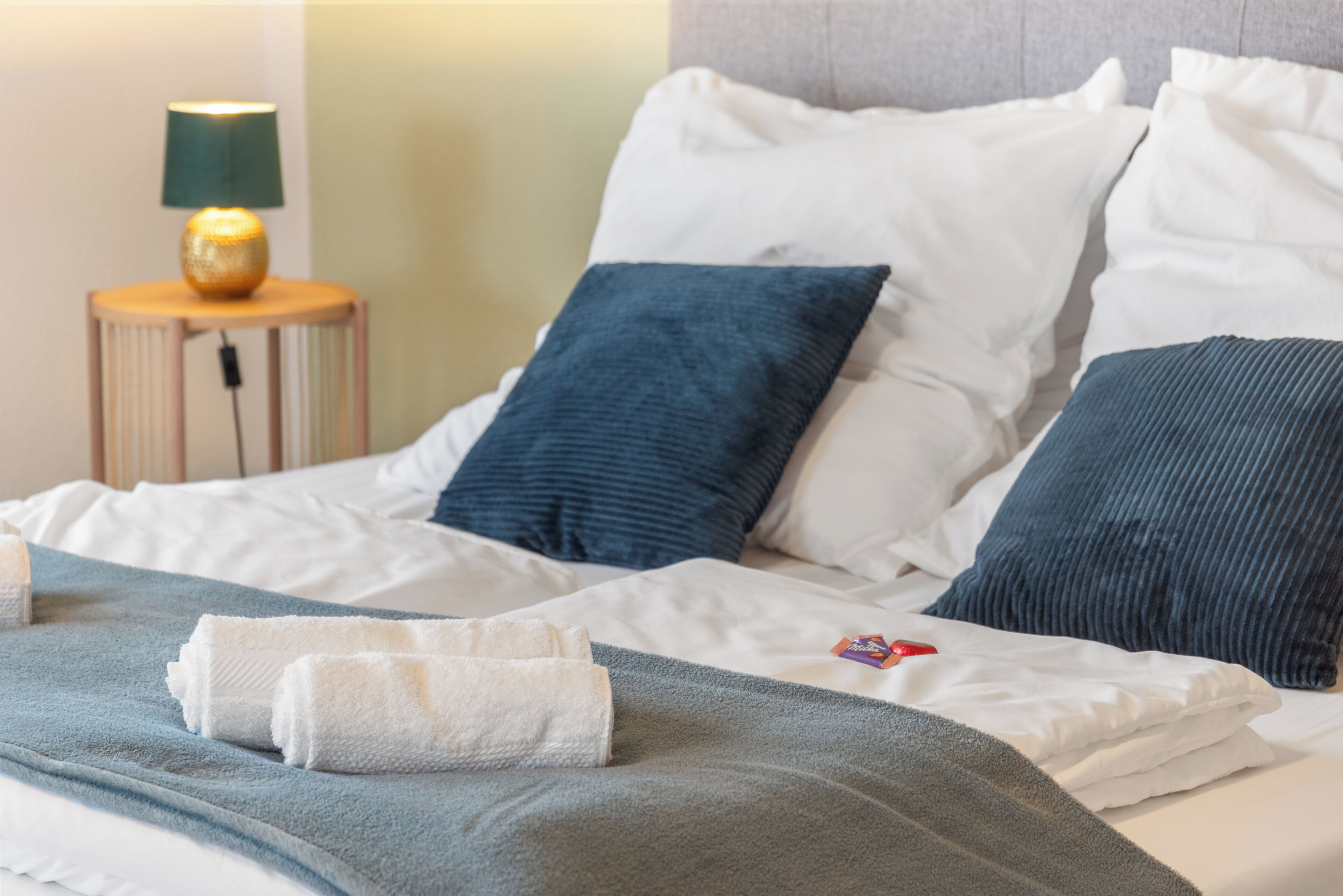 A neatly made bed with white and blue bedding, a small round nightstand with a lamp, and a wall with a textured pattern.