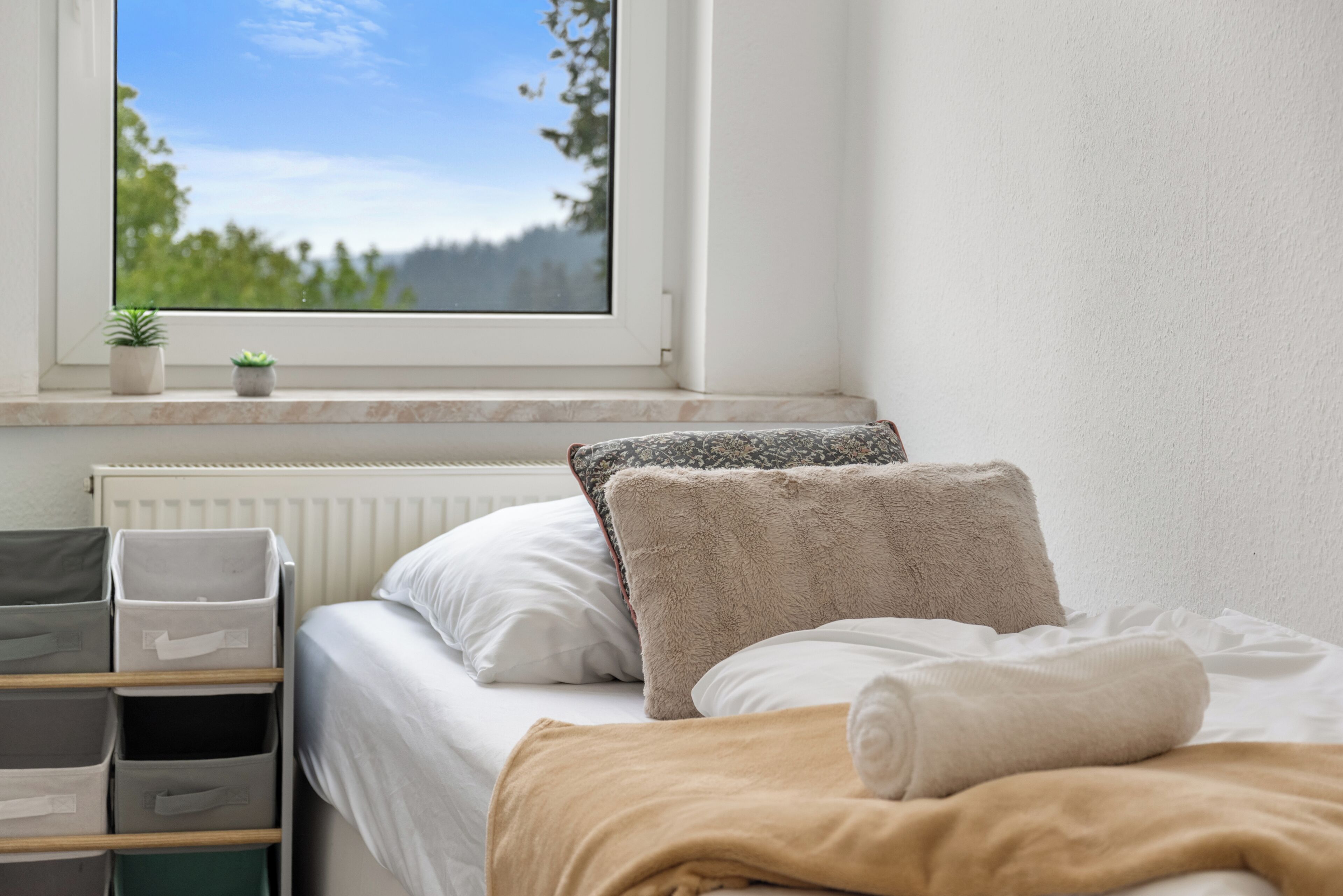A bedroom with a bed, pillows, a window with a view of trees, and storage bins.