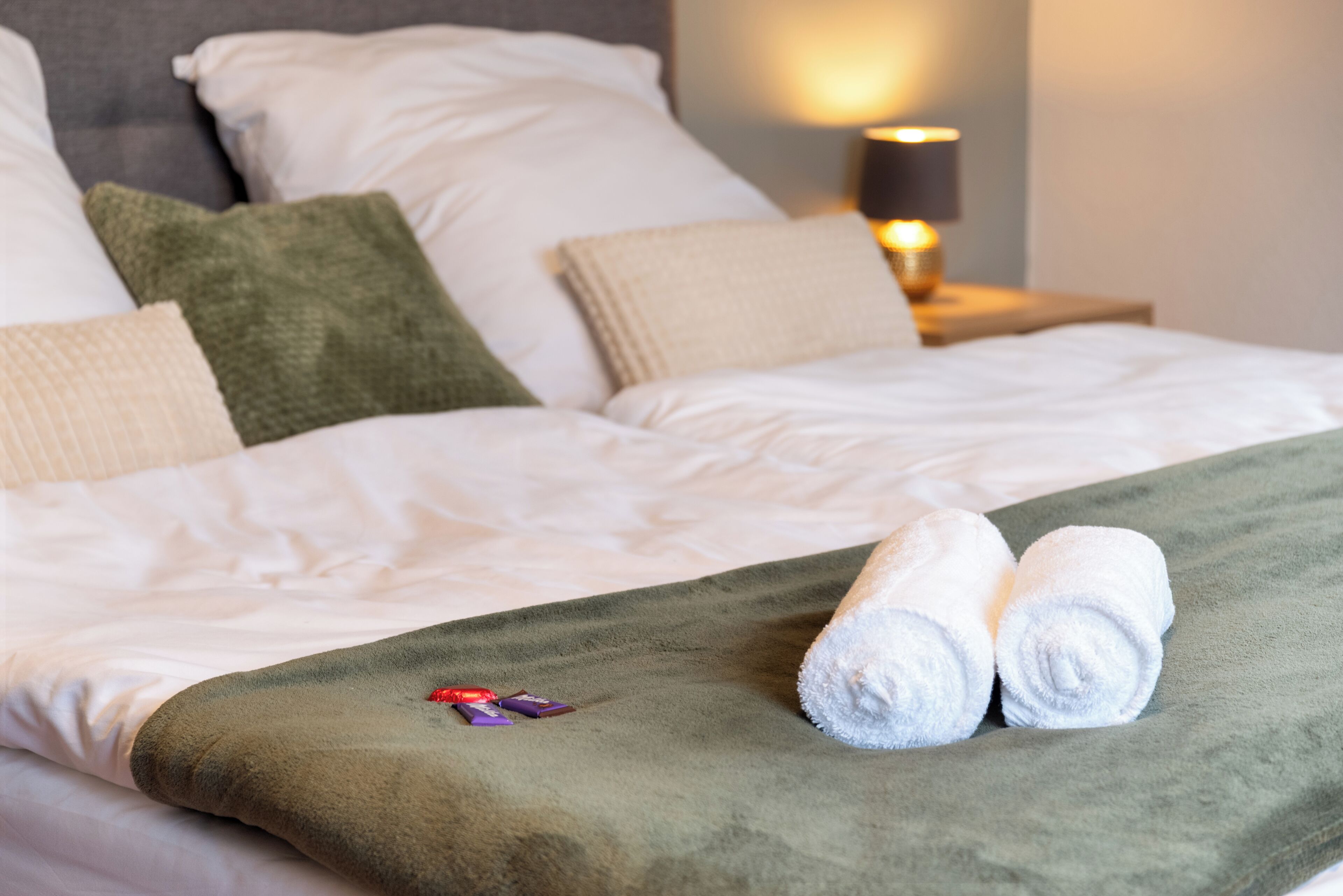 Una cama bien hecha con sábanas blancas, un plaid verde y dos toallas blancas enrolladas.