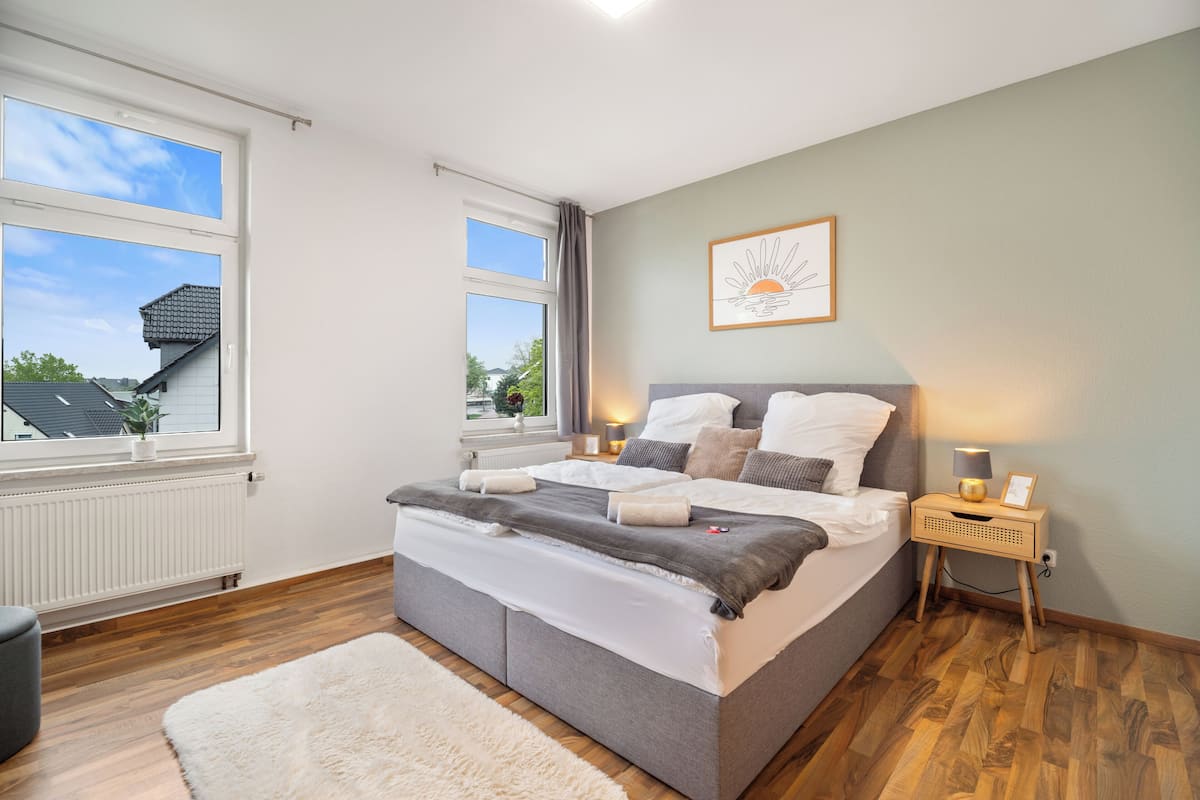 A bedroom with a bed, bedside tables, a window, a radiator, and a view of the outside.