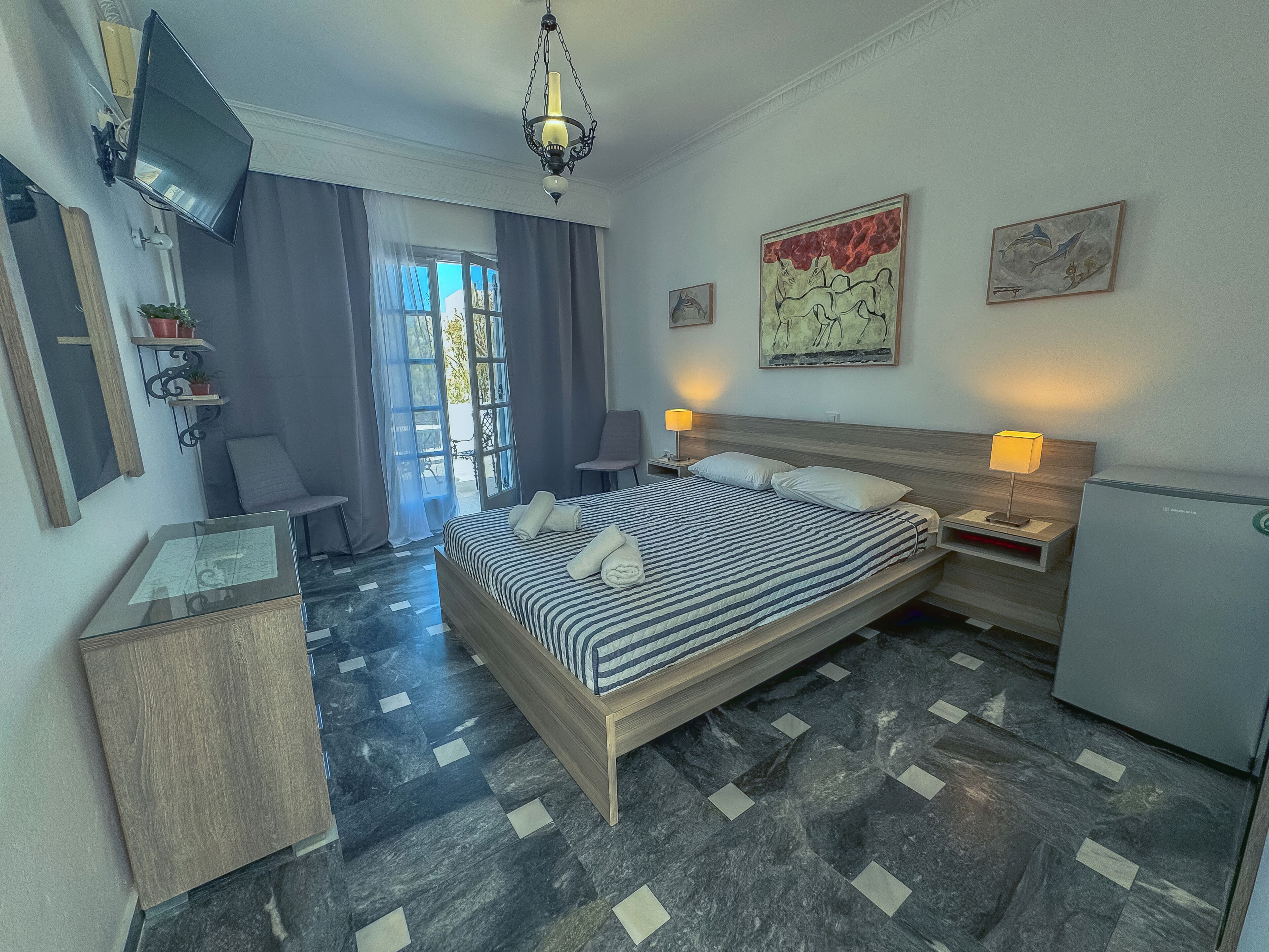 A hotel room with a bed, bedside lamps, a desk, a chair, and artwork on the walls.