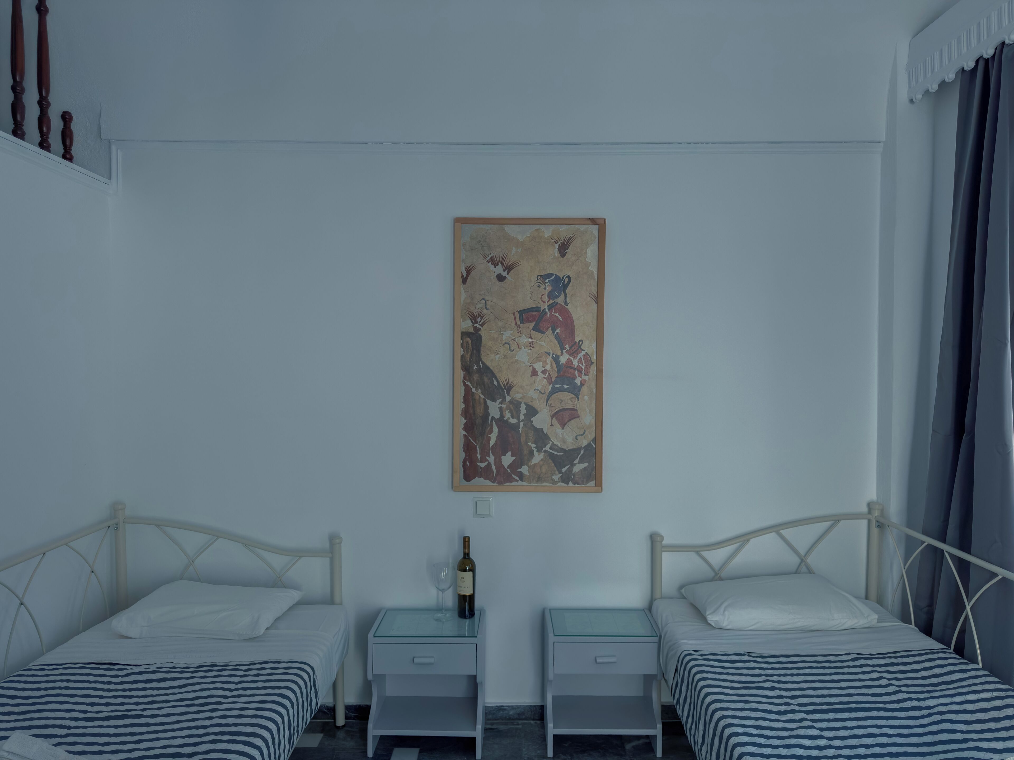 A bedroom with two single beds, a nightstand with a bottle and glass, and a wall-mounted painting.