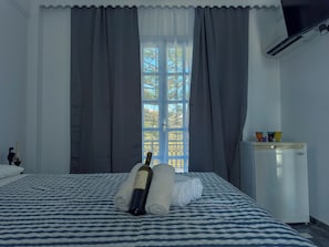 Habitación de hotel con cama, botella de vino, refrigerador pequeño y ventana con cortinas.