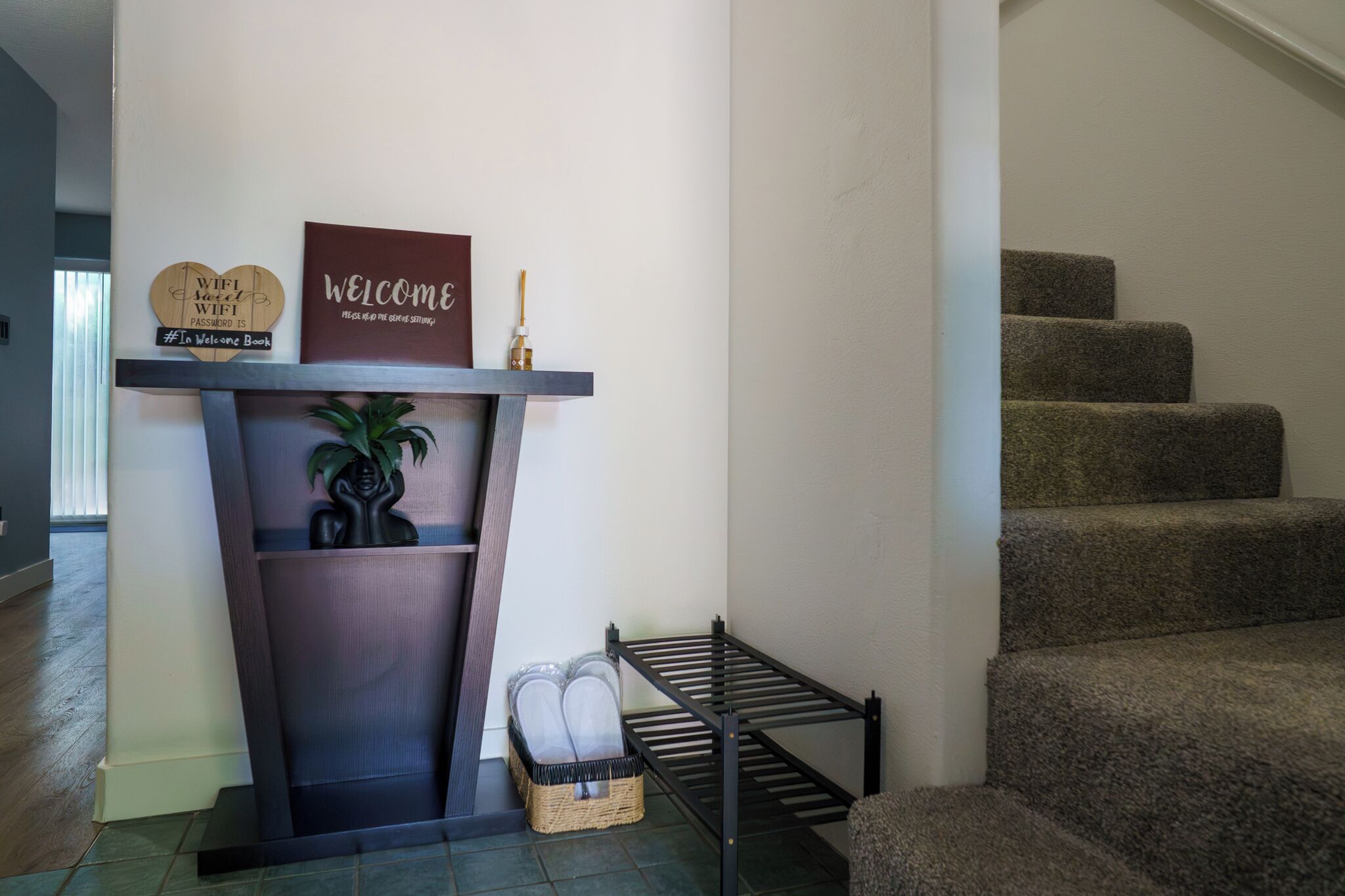 Un escalier avec des marches recouvertes de moquette, un panneau d’accueil, une petite étagère avec une plante et un porte-objets fixé au mur.