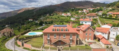 A large, red-roofed building with a central staircase, surrounded by smaller houses, set in a hilly area with a clear sky.