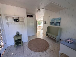 A room with a door, a small table with a jar, a bench, a rug, and a painting on the wall.