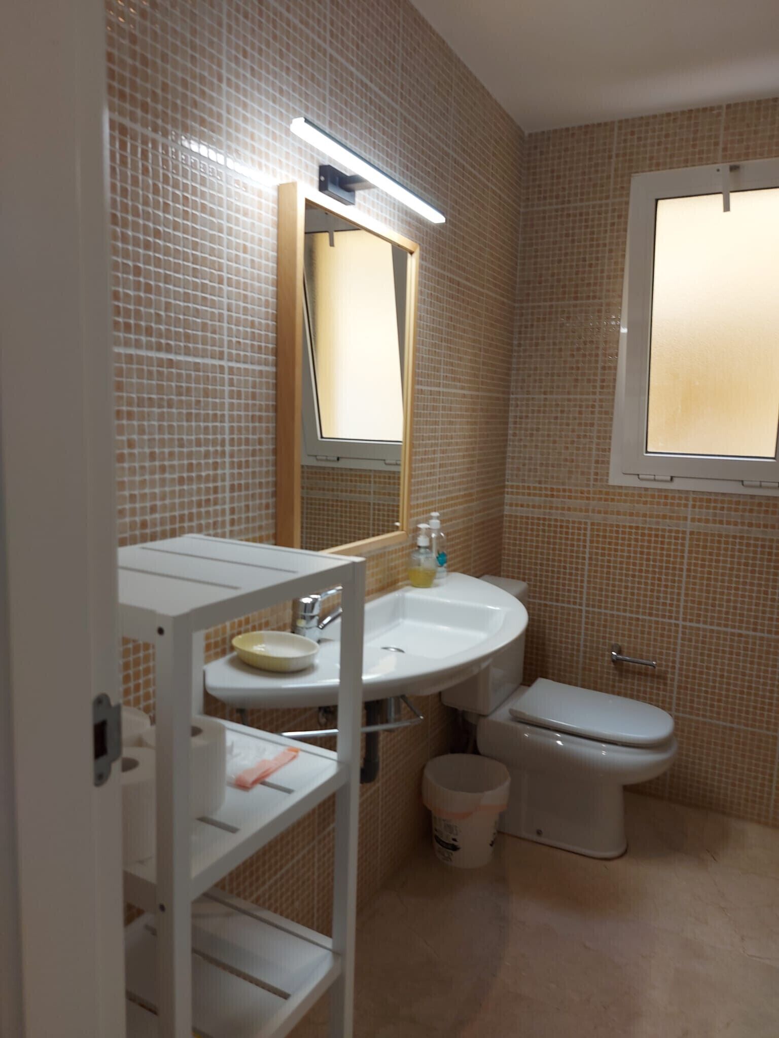 Une salle de bain avec un lavabo blanc, un miroir et des toilettes. Il y a une fenêtre au-dessus des toilettes et une étagère avec des articles de toilette.